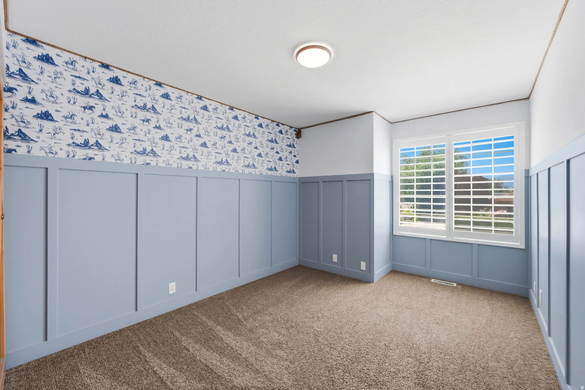 Carpeted room featuring light blue board and batten paneling, decorative wallpaper, a dual-pane window with white plantation shutters, and a flush-mount ceiling light fixture