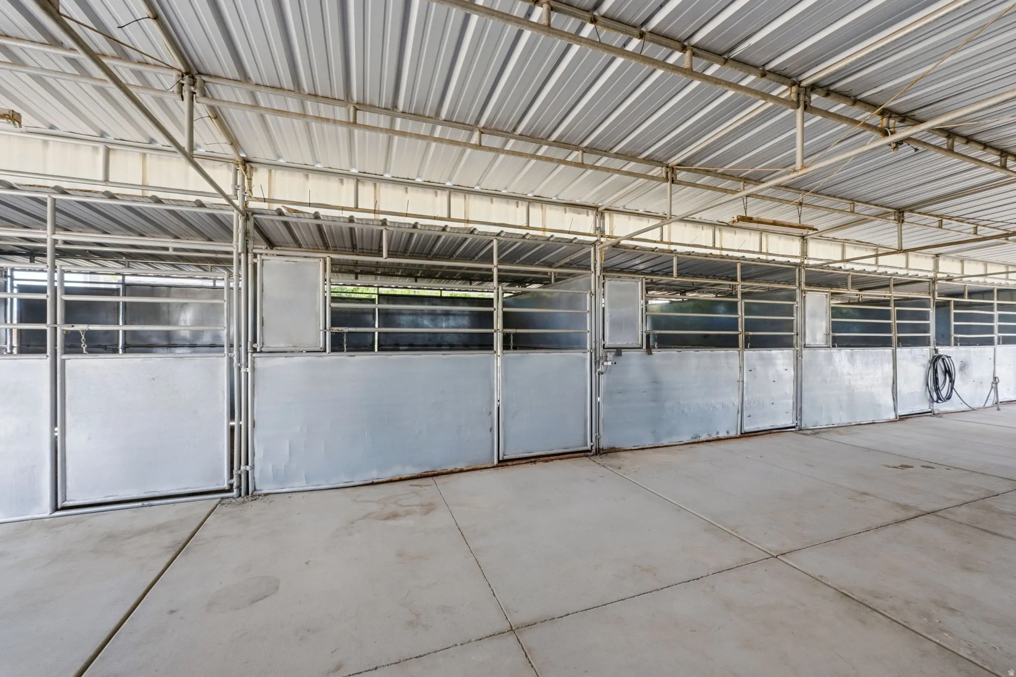 Covered barn structure featuring a corrugated metal roof, concrete flooring, and multiple metal stalls with gates