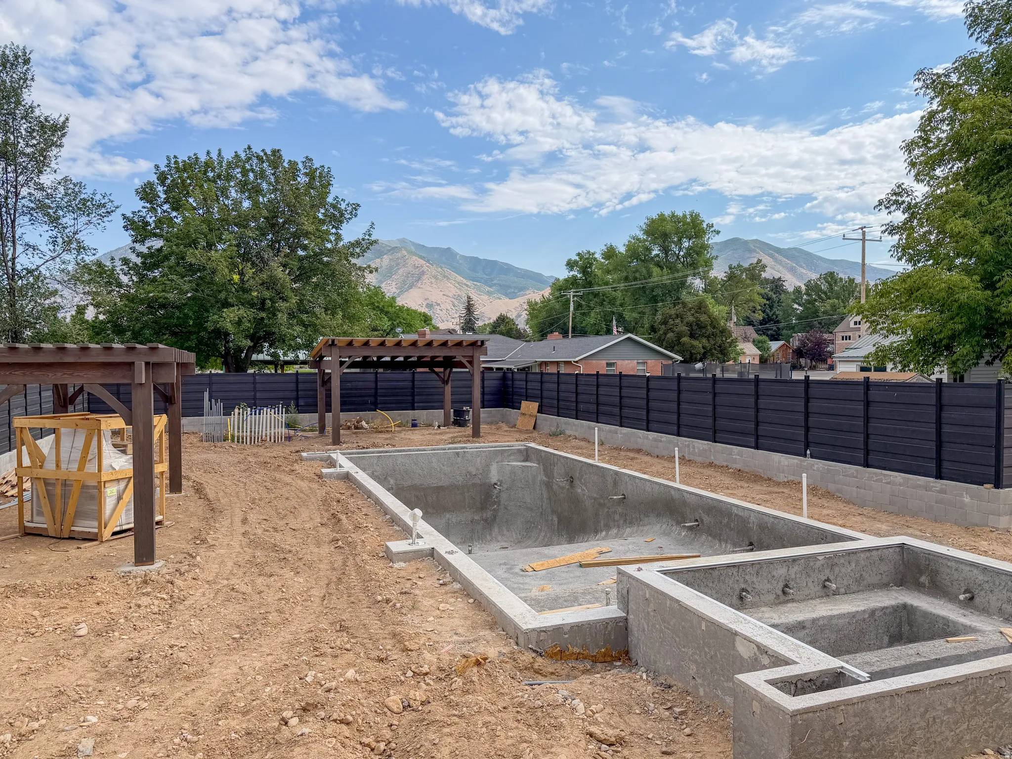 Backyard under construction featuring a concrete pool and spa structure, two wooden pergolas, and a dark privacy fence