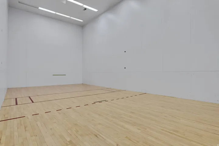 Dedicated indoor racquetball court featuring light wood flooring, white panel walls, and overhead fluorescent lighting