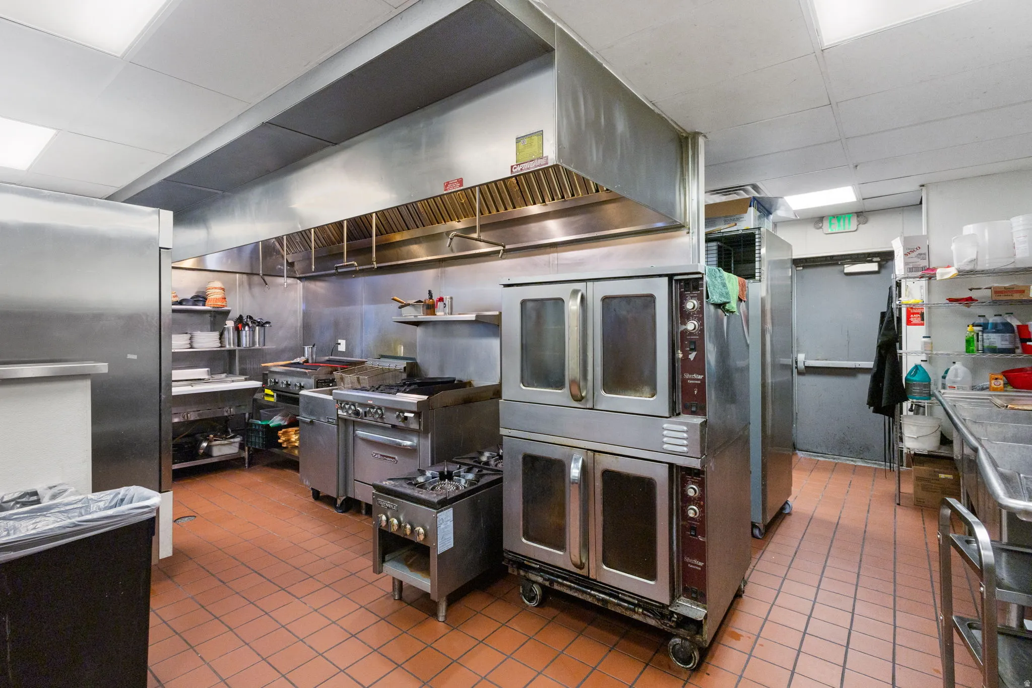 Commercial kitchen featuring stainless steel appliances, a large ventilation hood system, dual convection ovens, multiple ranges, and a three-compartment sink