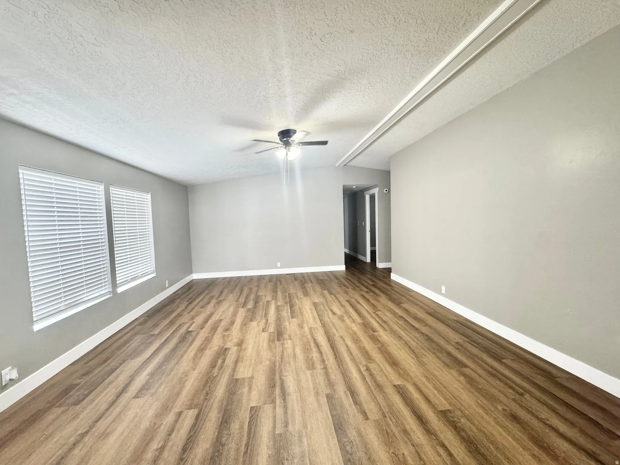 Spacious interior room featuring wood-finish flooring, two windows with horizontal blinds, a ceiling fan with integrated lighting, and neutral wall tones