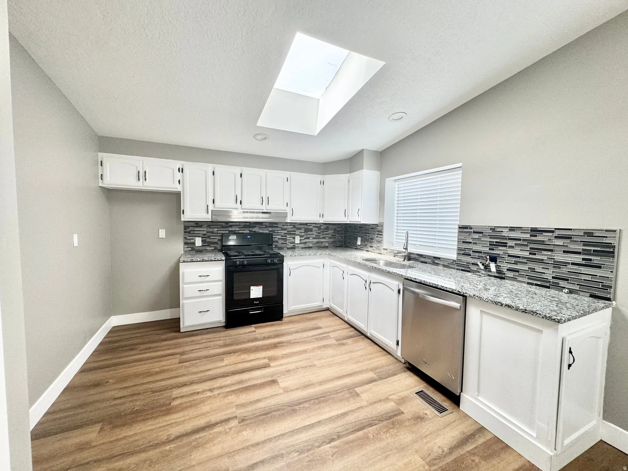 Kitchen featuring wood-finish flooring, white cabinetry, speckled countertops, a mosaic tile backsplash, and a skylight