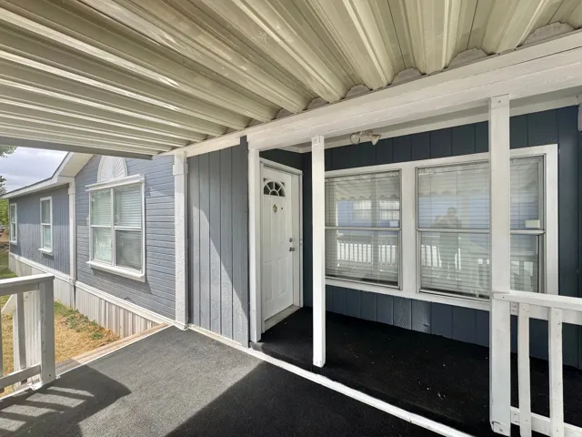 Covered porch entry featuring a white paneled door with decorative glass, light blue siding, white window trim, and a grey outdoor carpet