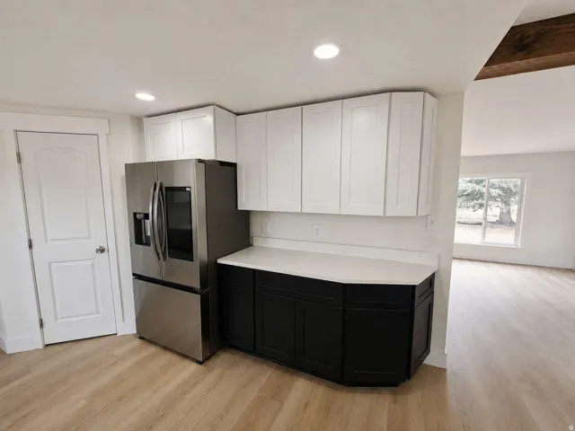 Stainless steel refrigerator, two-tone cabinetry with white upper and black lower units, light-toned wood-finish flooring, and recessed lighting