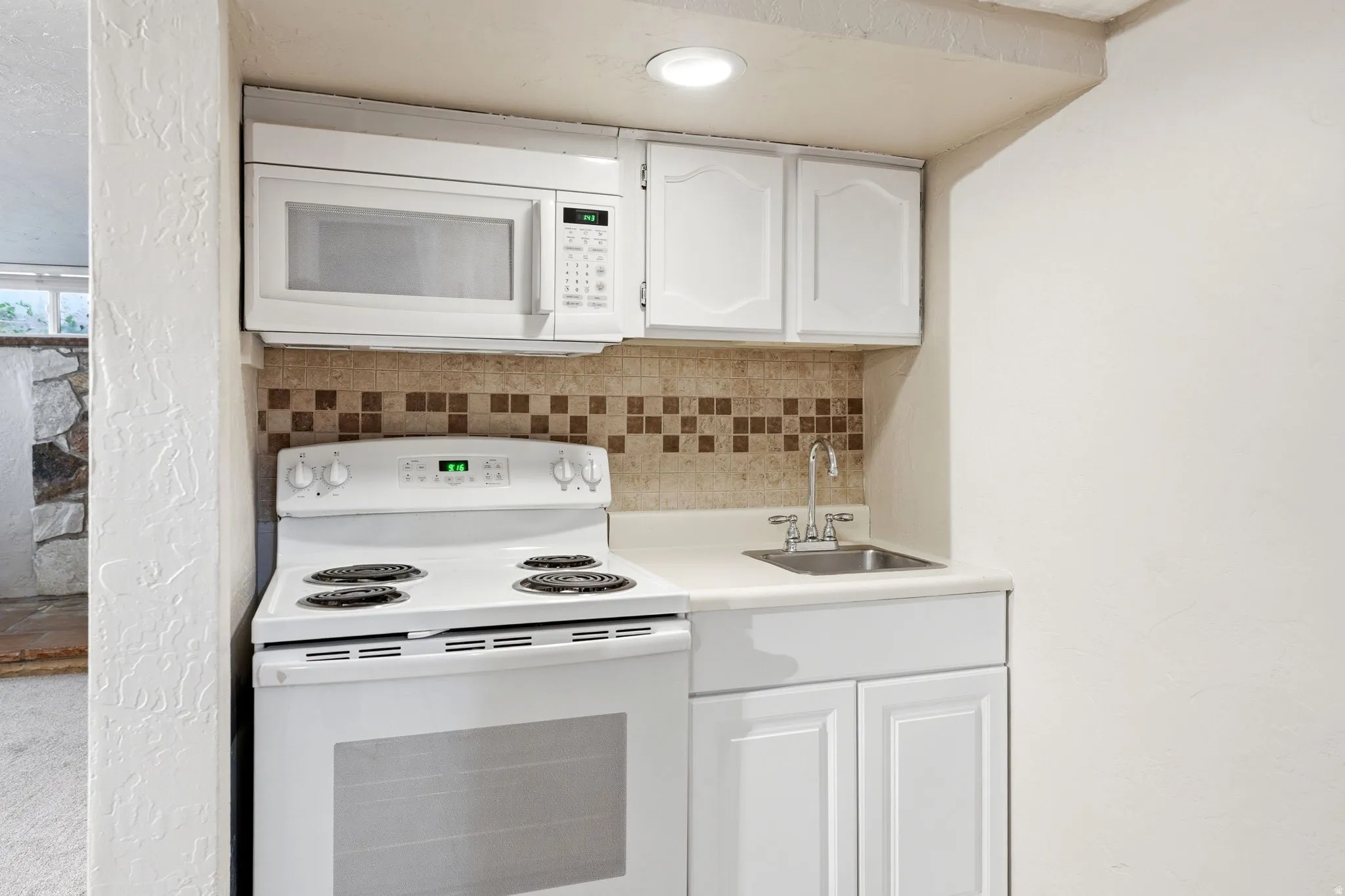 Compact kitchen area featuring white appliances, a white laminate countertop, a stainless steel sink with a chrome faucet, and a brown and beige mosaic tile backsplash