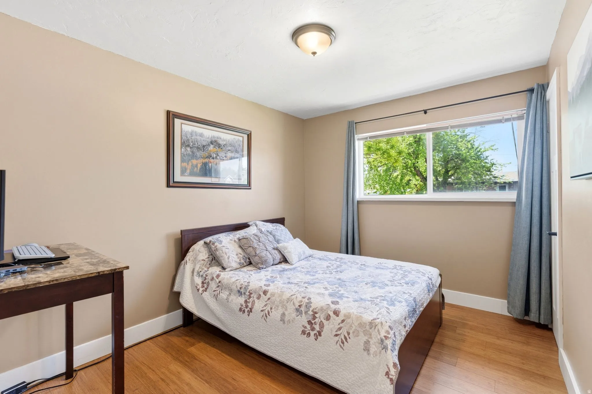 Bright room featuring a large window, warm wood-finish flooring, neutral wall tones, and a ceiling-mounted light fixture