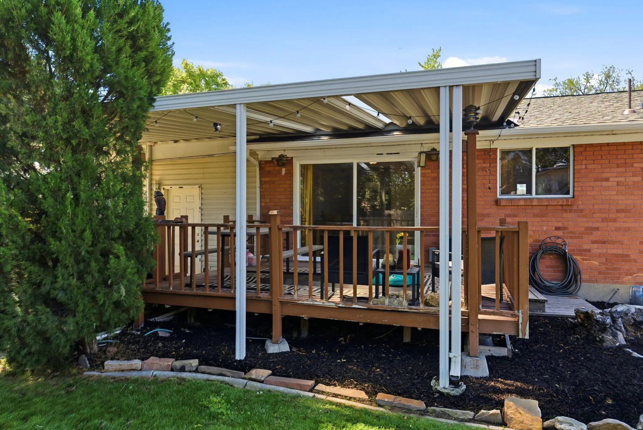 Covered wood-plank deck with railing, featuring a sliding glass door and brick exterior