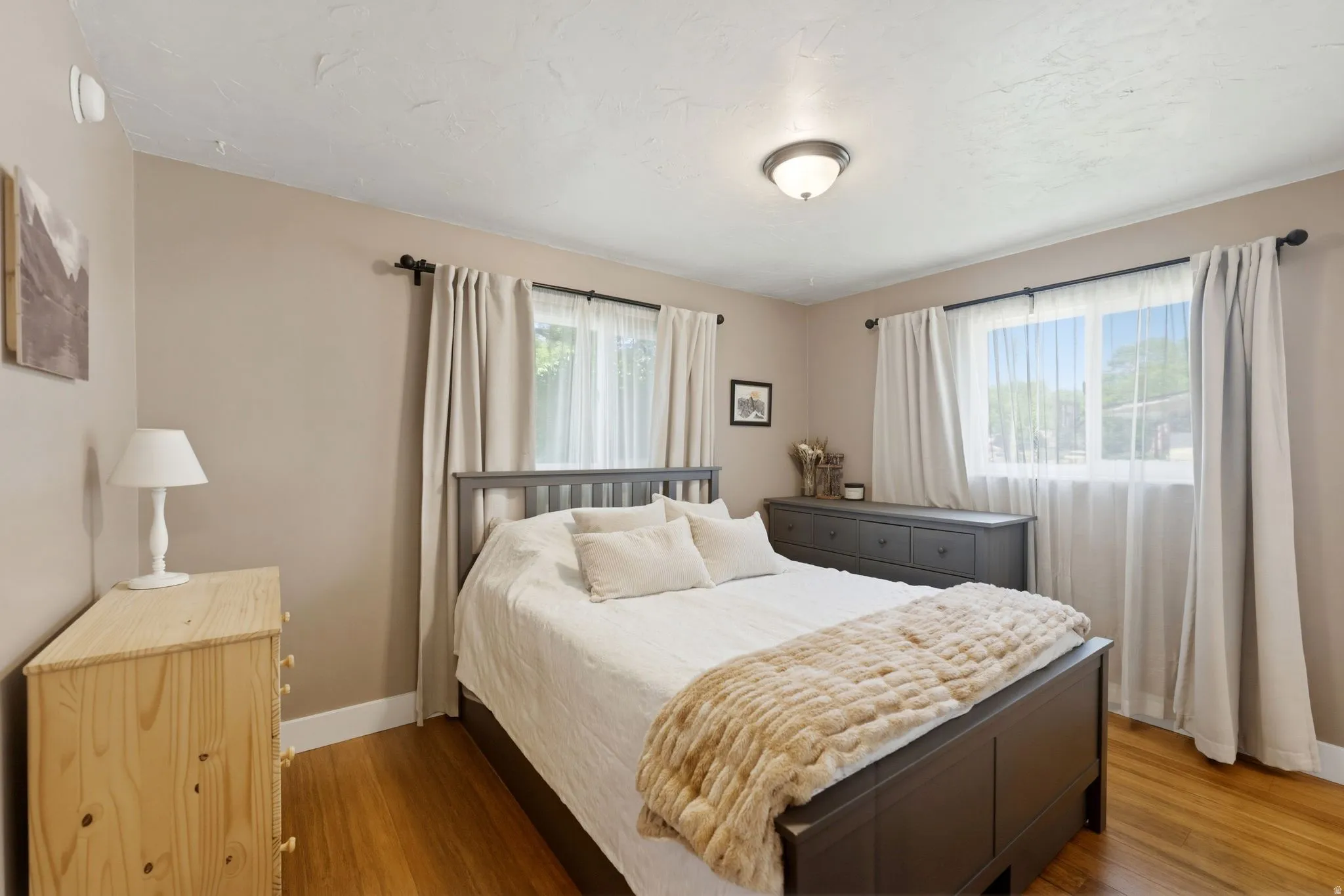 Bright bedroom featuring two windows, wood-finish flooring, neutral wall tones, and a ceiling-mounted light fixture