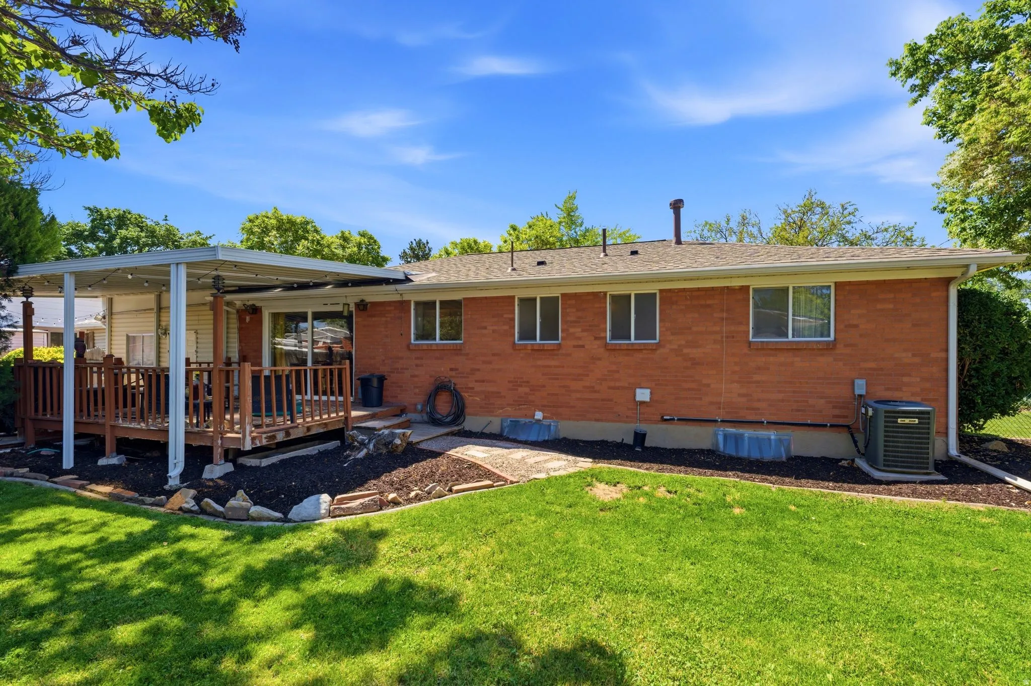 Rear exterior featuring brick siding, a covered wood deck, sliding glass door access, multiple windows, and a landscaped garden bed