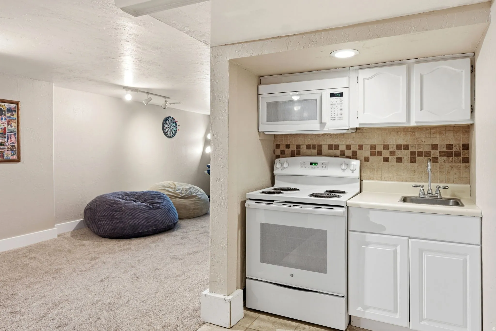 Compact kitchen area featuring a white electric range, over-the-range microwave, stainless steel sink, tile backsplash, and white cabinetry
