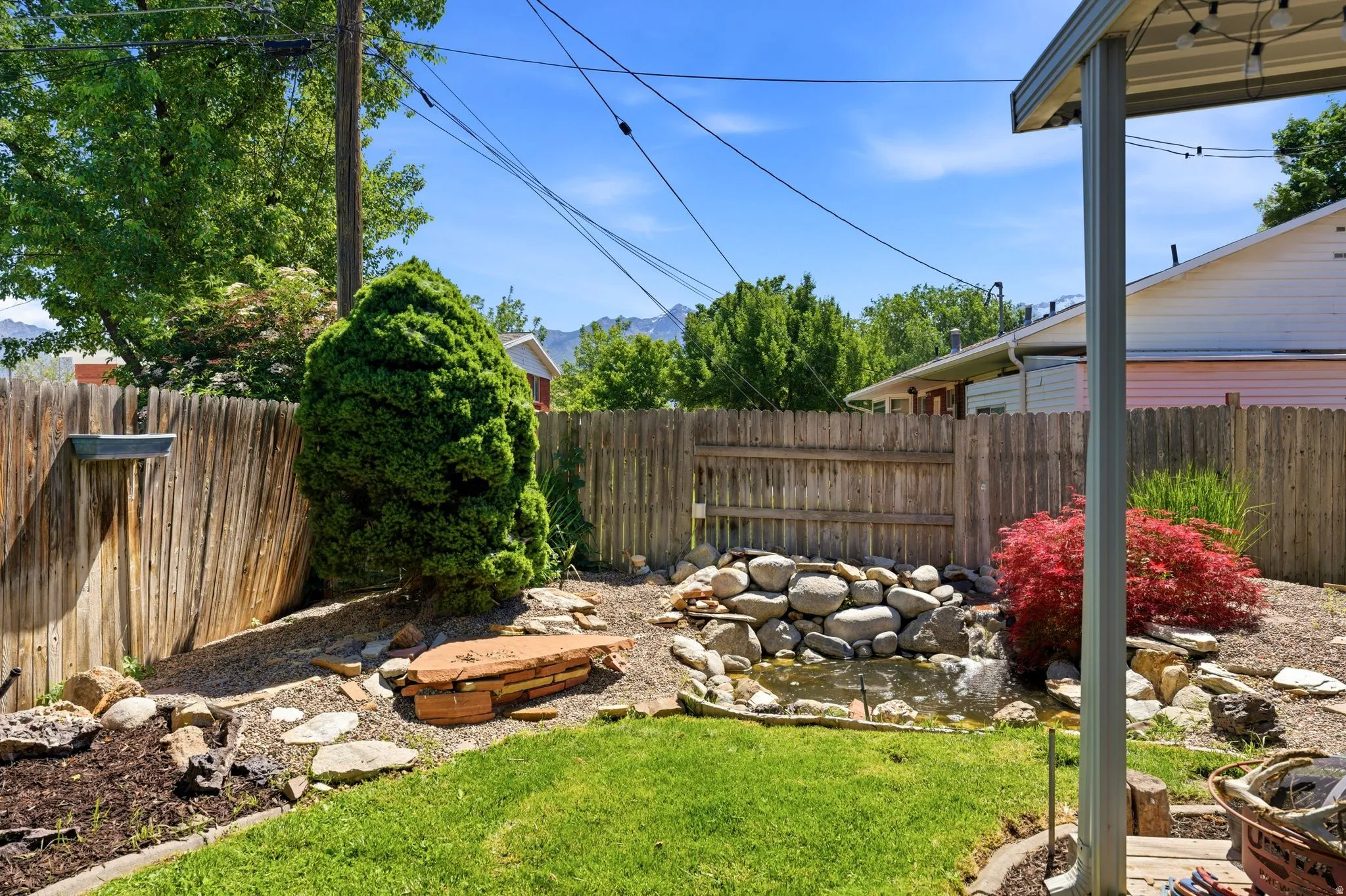 Expansive backyard featuring a rock-lined water feature, established landscaping, wooden privacy fencing, and a covered patio area
