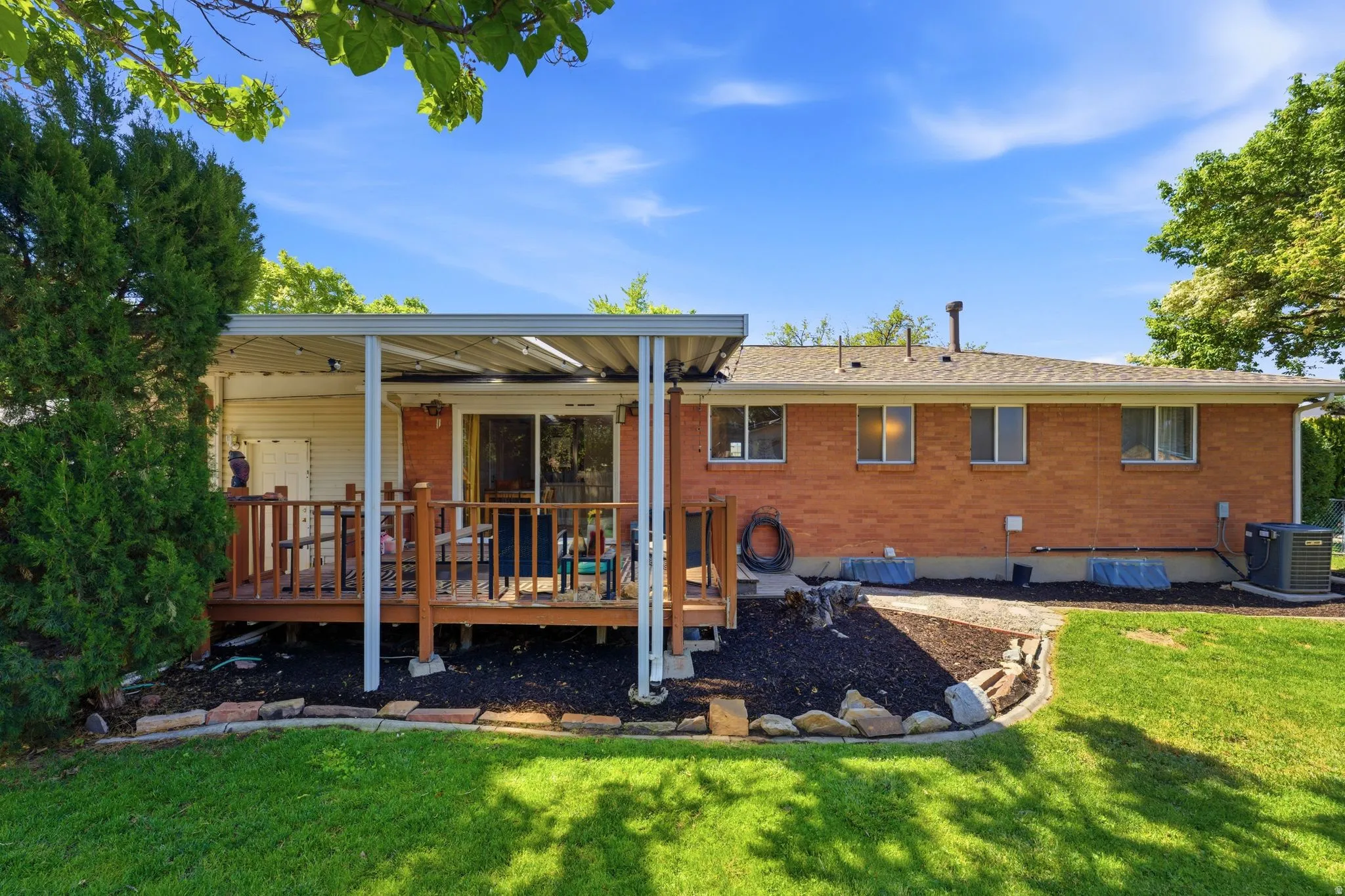 Expansive backyard featuring a covered deck with railings, a manicured lawn, established landscaping, and a brick exterior