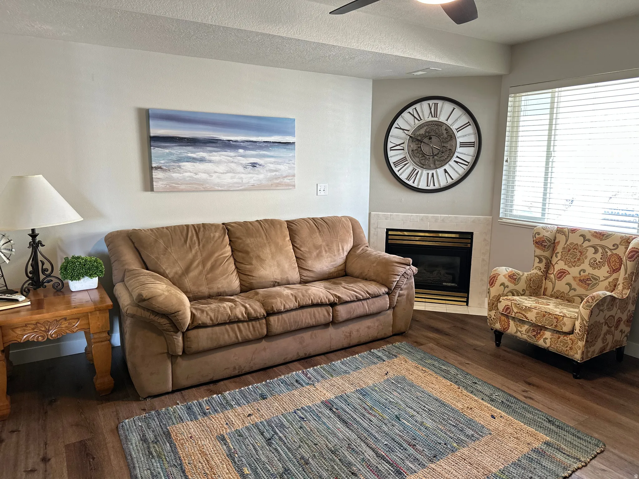 Living area featuring wood-finish flooring, a corner fireplace with a light-toned surround, a ceiling fan, and window blinds