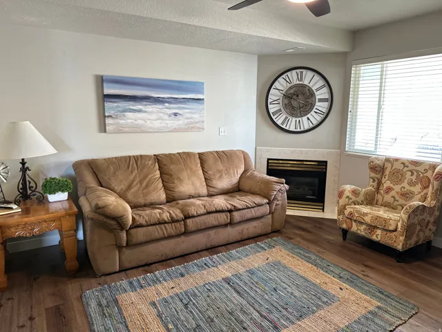Living area featuring wood-finish flooring, a corner fireplace with a light-toned surround, a ceiling fan, and window blinds