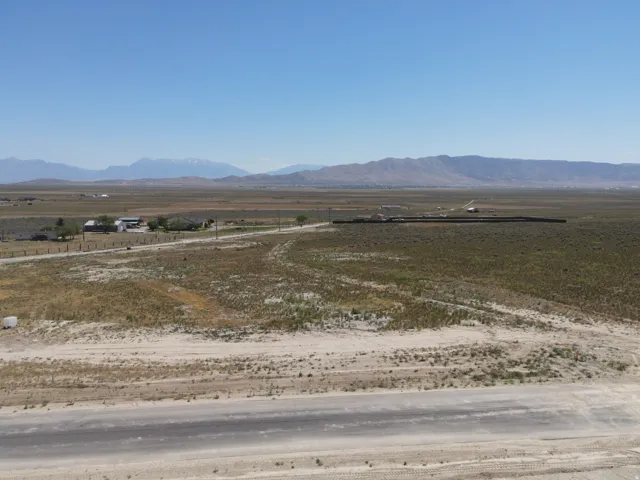 Expansive undeveloped land featuring a dirt road, distant residential structures, and a mountain backdrop under a clear sky
