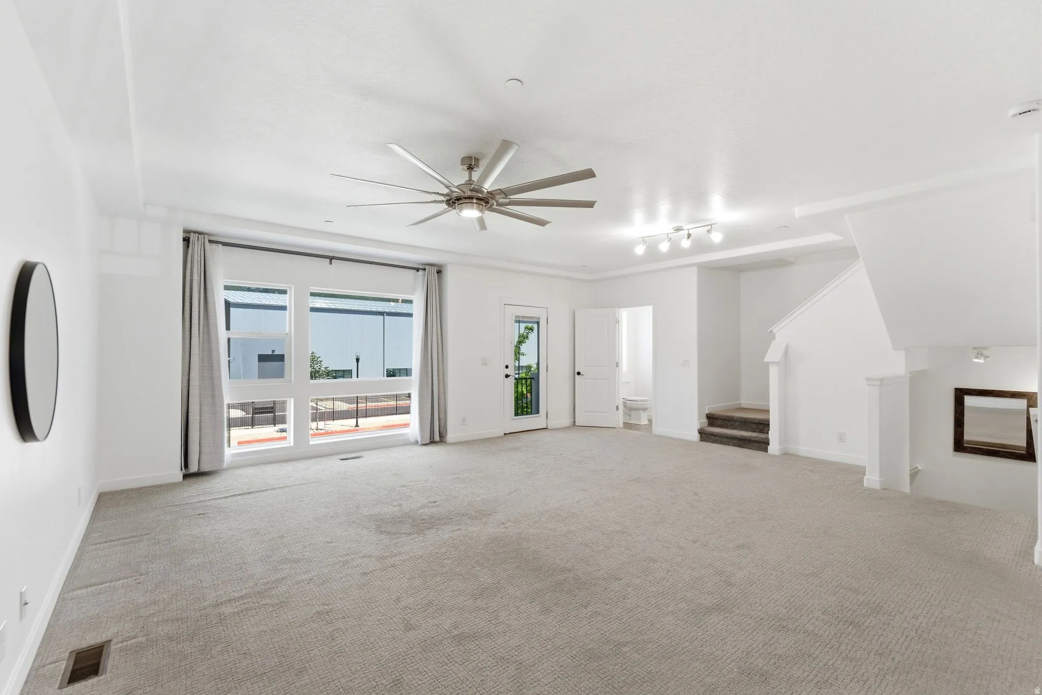 Expansive living area featuring light gray carpeting, a contemporary ceiling fan, and large windows with gray drapery