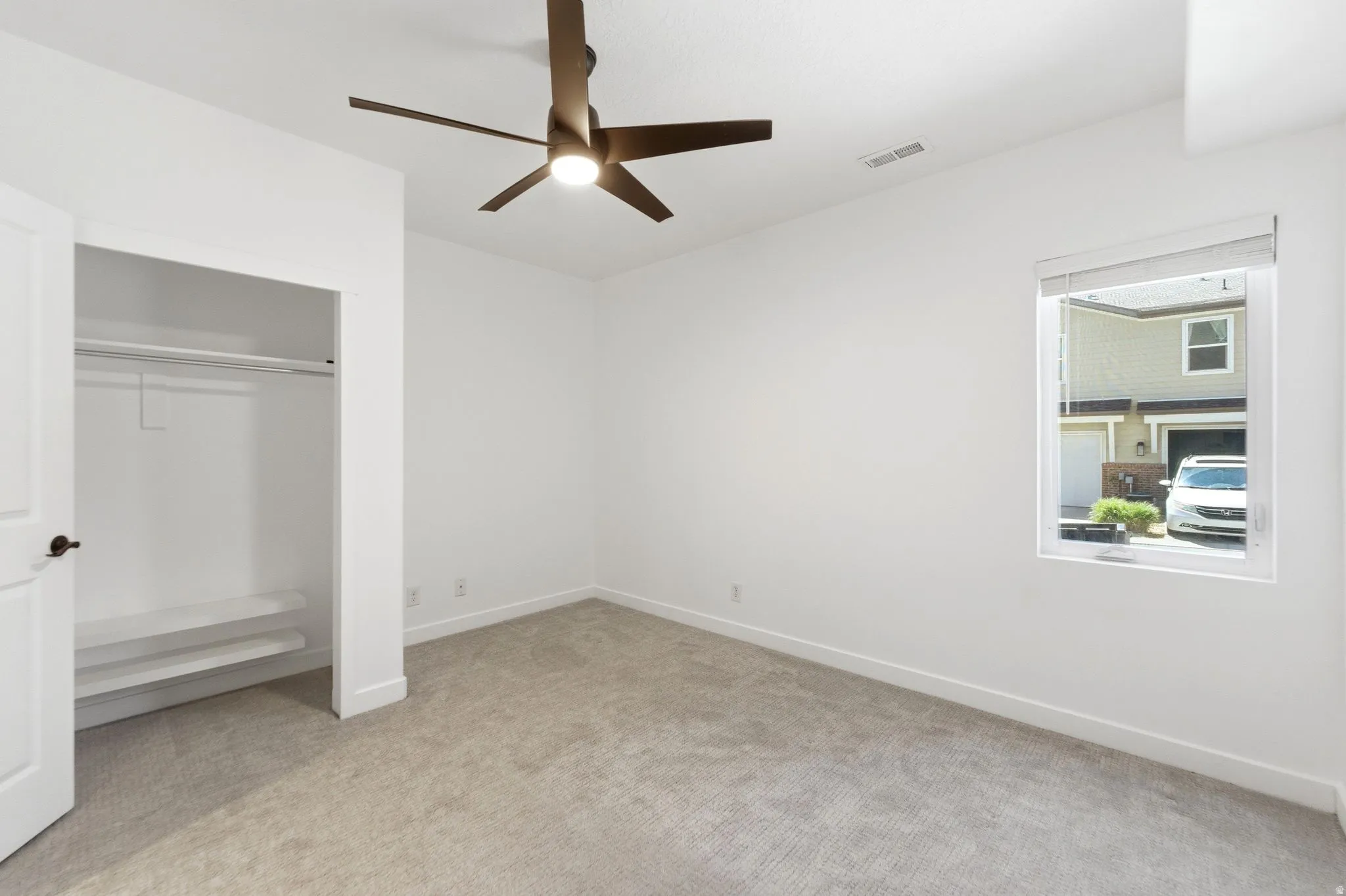 Bright room featuring neutral carpeting, white walls, a modern ceiling fan with lighting, a window with roller blinds, and an open closet with built-in shelving and a clothing rod