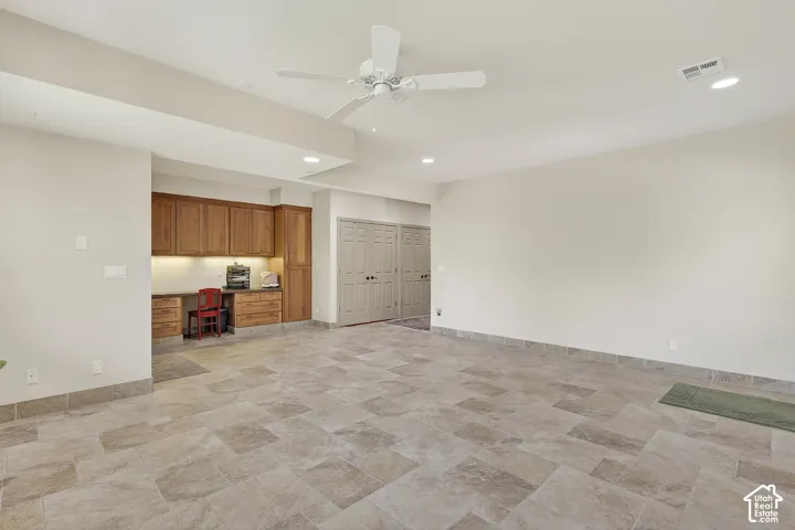 Spacious room featuring large format tile flooring, a built-in wood-finish desk with upper and lower cabinetry, recessed lighting, and a ceiling fan