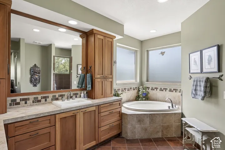 Bathroom featuring a corner soaking tub with surrounding tiled deck, dual windows with frosted glass, wood-finish vanity with an integrated sink, patterned mosaic tile backsplash, and recessed lighting