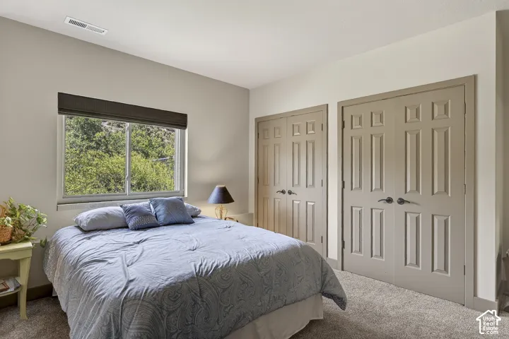 Bedroom featuring a window with a dark roller shade, two paneled closet doors with dark hardware, a ceiling-mounted HVAC vent, and neutral-toned carpet flooring