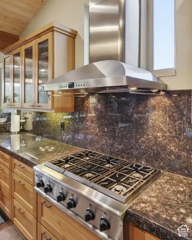 Stainless steel gas range with a matching range hood, granite countertops, granite backsplash, and light wood cabinetry with glass-front upper cabinets