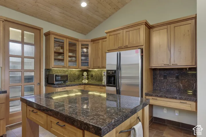Kitchen featuring a large island with a dark stone countertop, light wood cabinetry, stainless steel refrigerator, and a wood-paneled vaulted ceiling