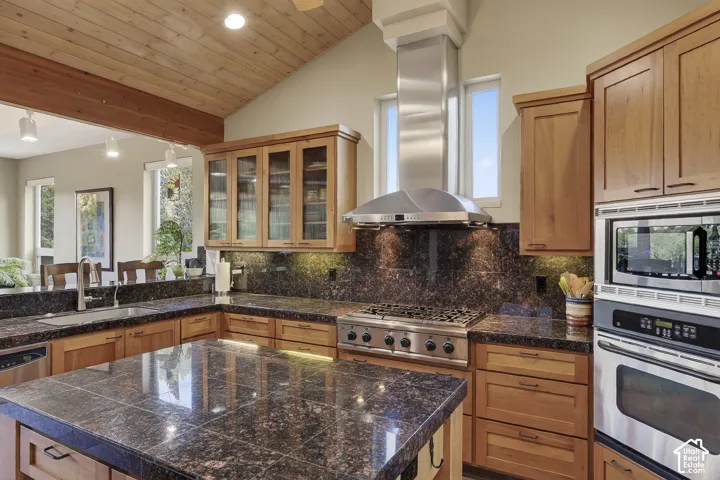 Kitchen featuring a vaulted wood-paneled ceiling, stainless steel range hood, built-in stainless steel appliances, dark stone countertops, and wood cabinetry with glass-front uppers