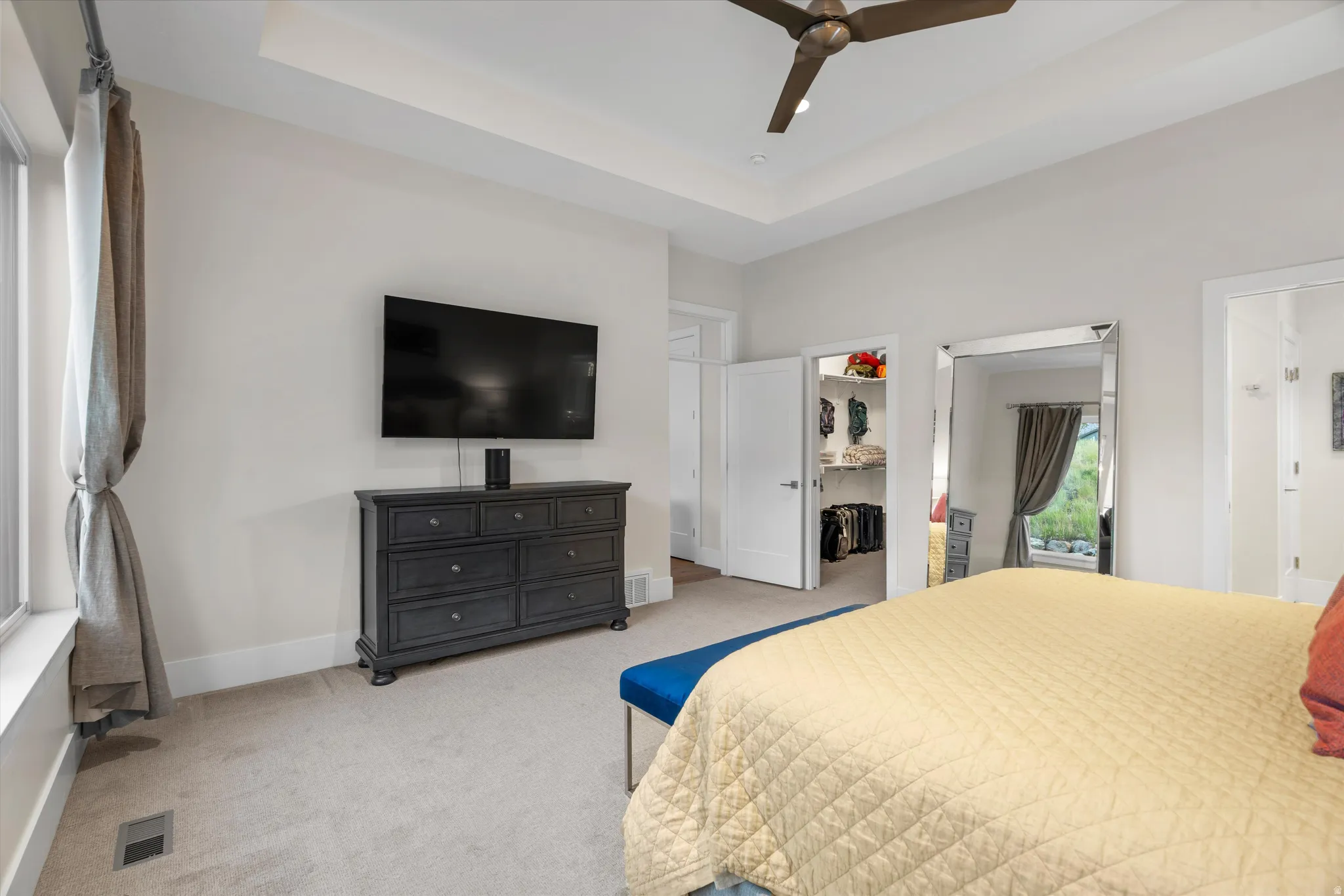 Tray ceiling with a ceiling fan, plush carpet flooring, large window with drapes, and an open closet featuring built-in shelving
