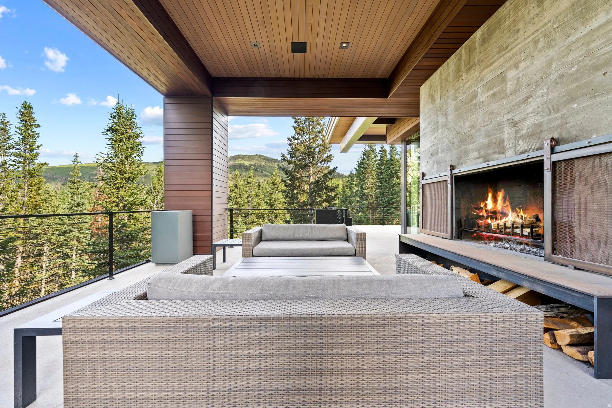 Spacious covered patio featuring a built-in outdoor fireplace with sliding metal screens, a wood-slat ceiling, and a modern glass railing system