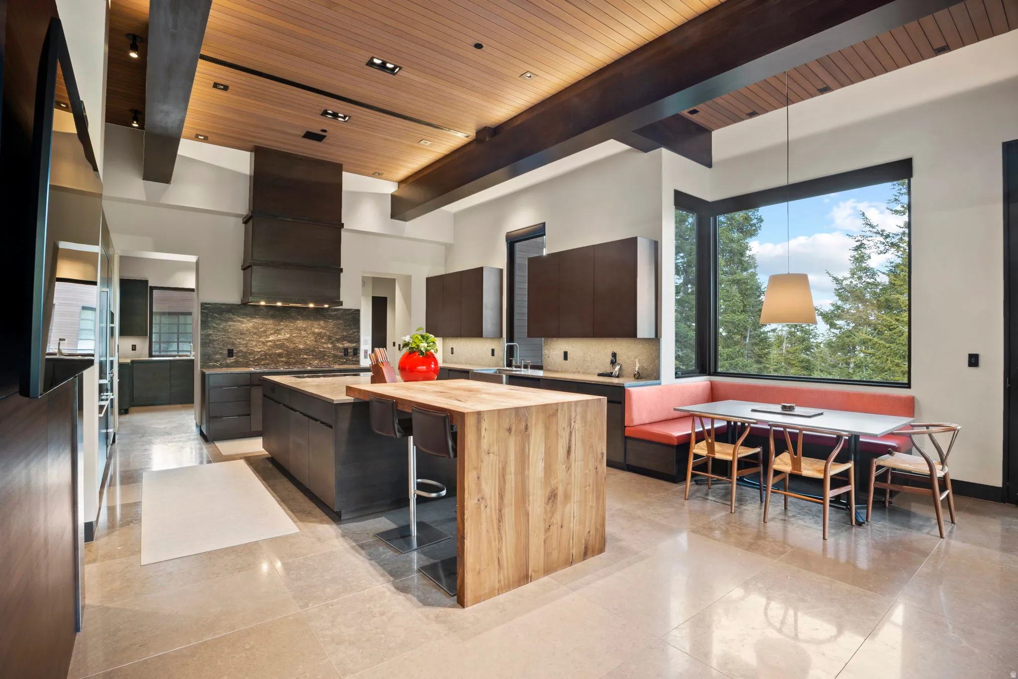 Contemporary kitchen featuring a large central island with a wood-finish breakfast bar, dark cabinetry, a stone-tiled backsplash, and a built-in banquette seating area under expansive corner windows
