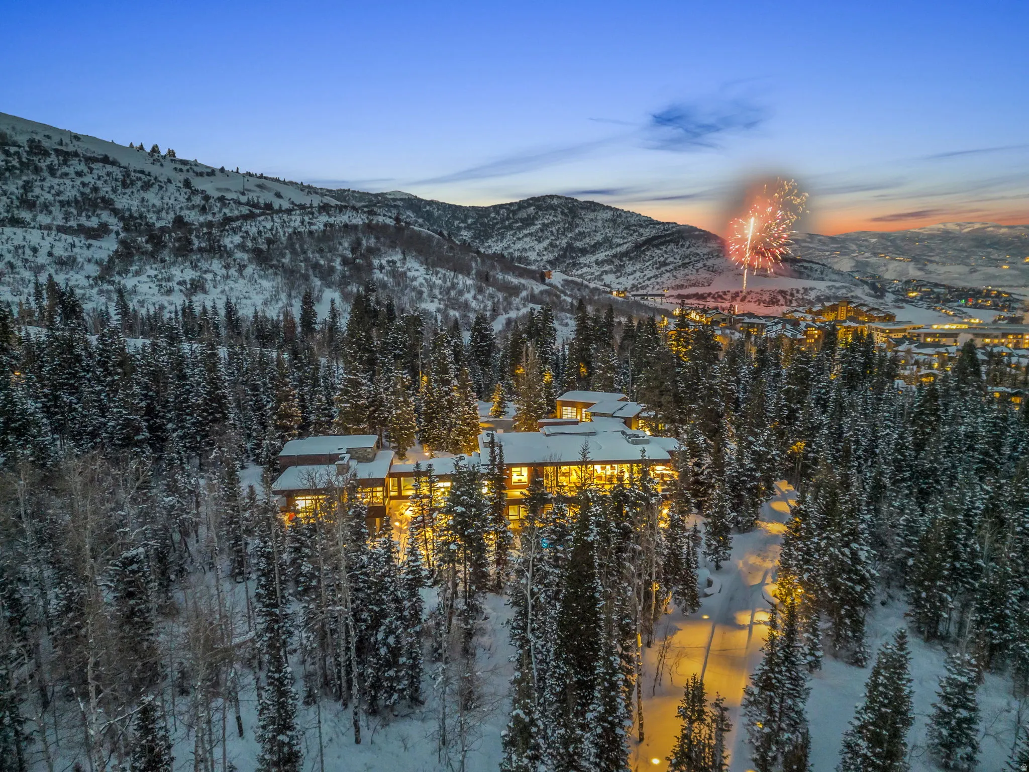 Extensive property nestled among snow-covered evergreen trees, featuring multiple levels, numerous illuminated windows, and a multi-peaked roofline