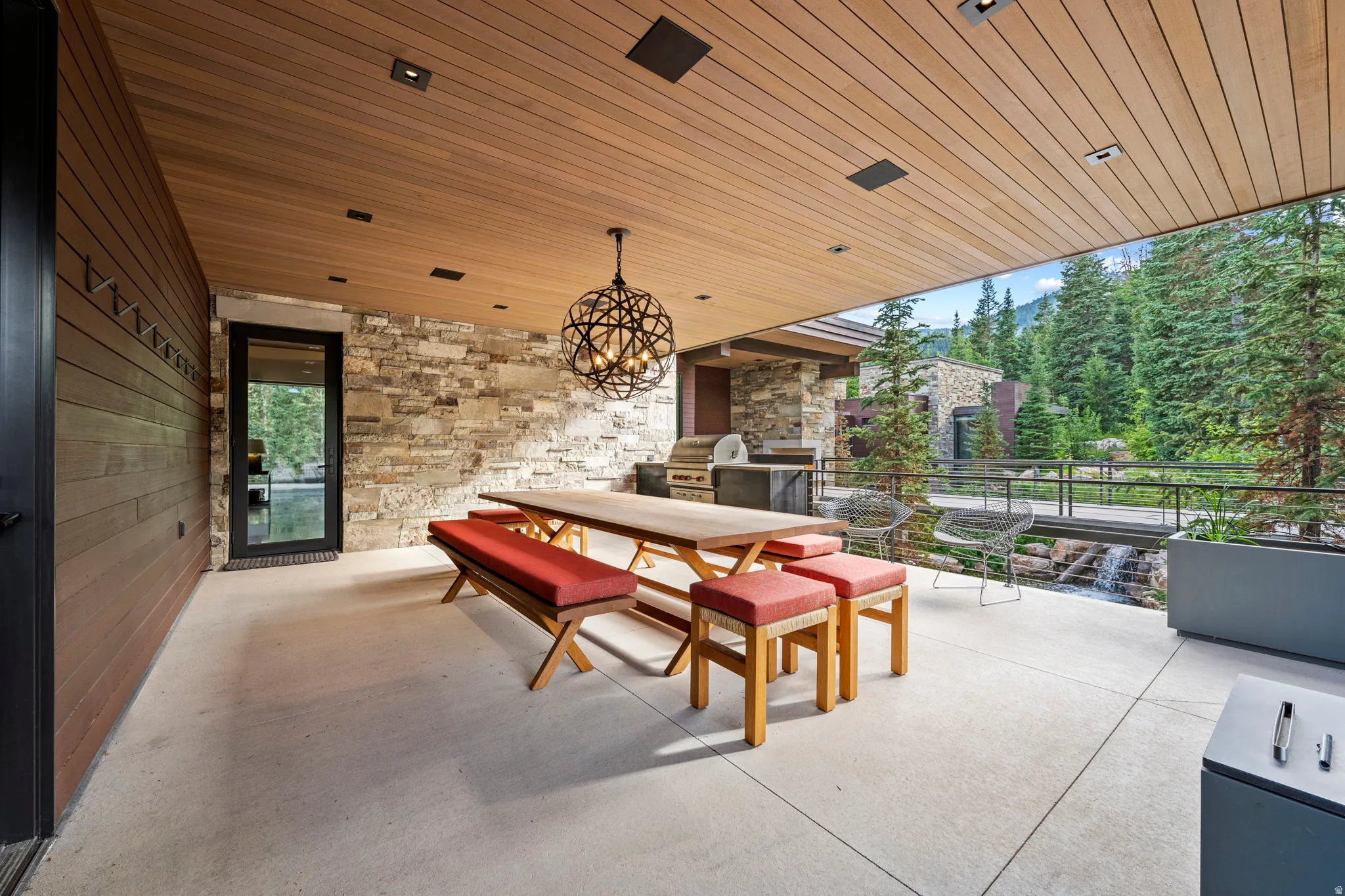Covered outdoor patio featuring a wood-finish ceiling with recessed lighting, a stone accent wall, an outdoor kitchen with built-in appliances, and a metal railing