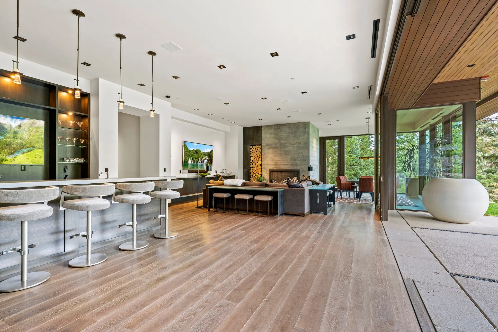 Open-concept interior featuring wood-finish flooring, a built-in bar with pendant lighting, a stone-clad fireplace, and expansive glass sliding doors