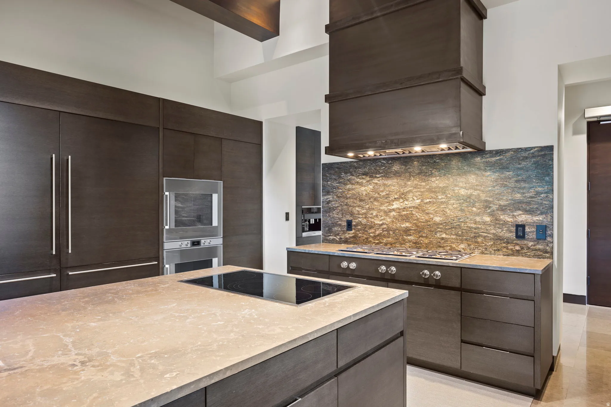 Kitchen featuring a large island with an integrated cooktop, dark wood-finish cabinetry, built-in stainless steel appliances, a stone backsplash, and a custom range hood