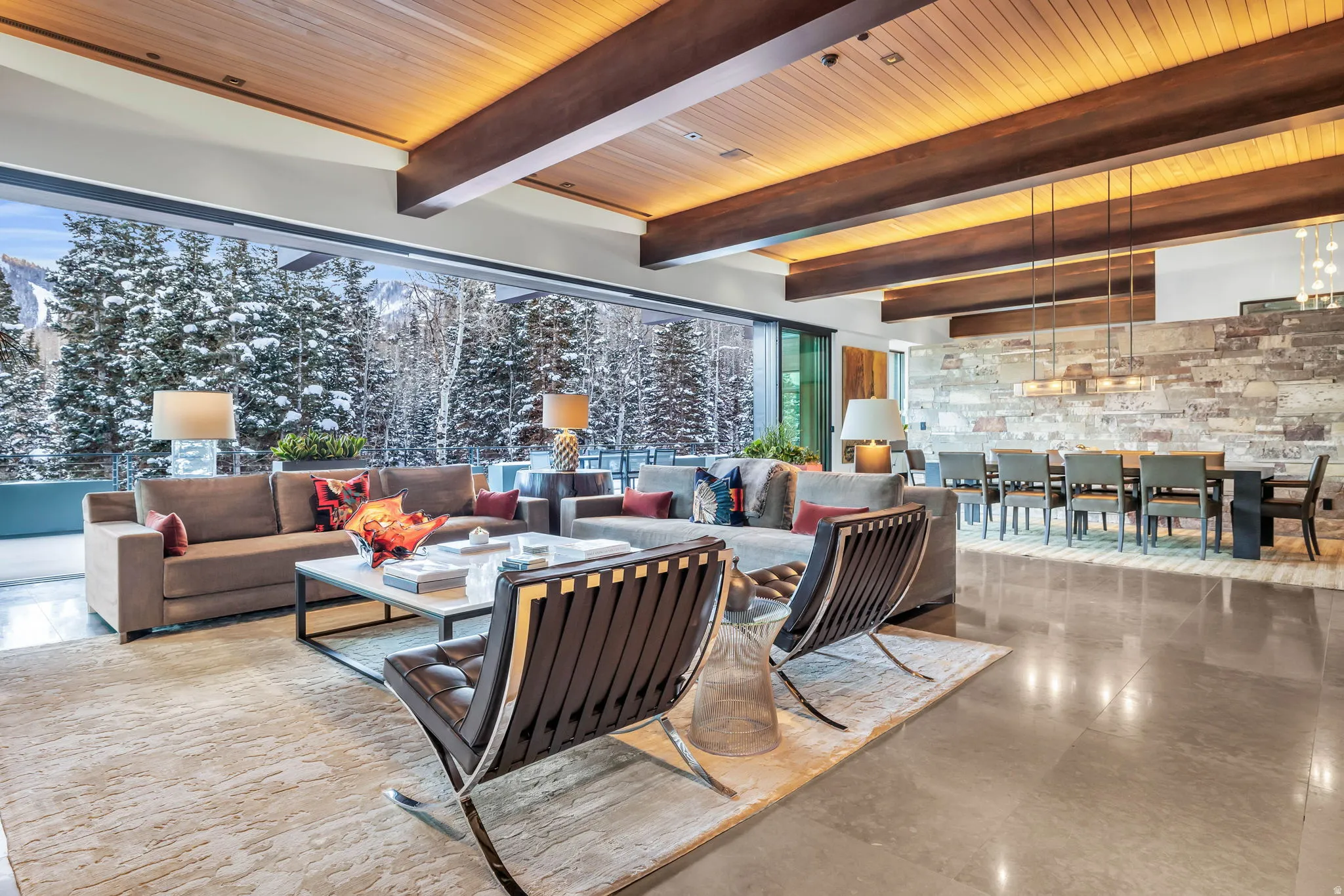Spacious open-concept living area featuring exposed wood beams, a full-height stone accent wall, and expansive glass doors opening to a snow-covered mountain and forest landscape