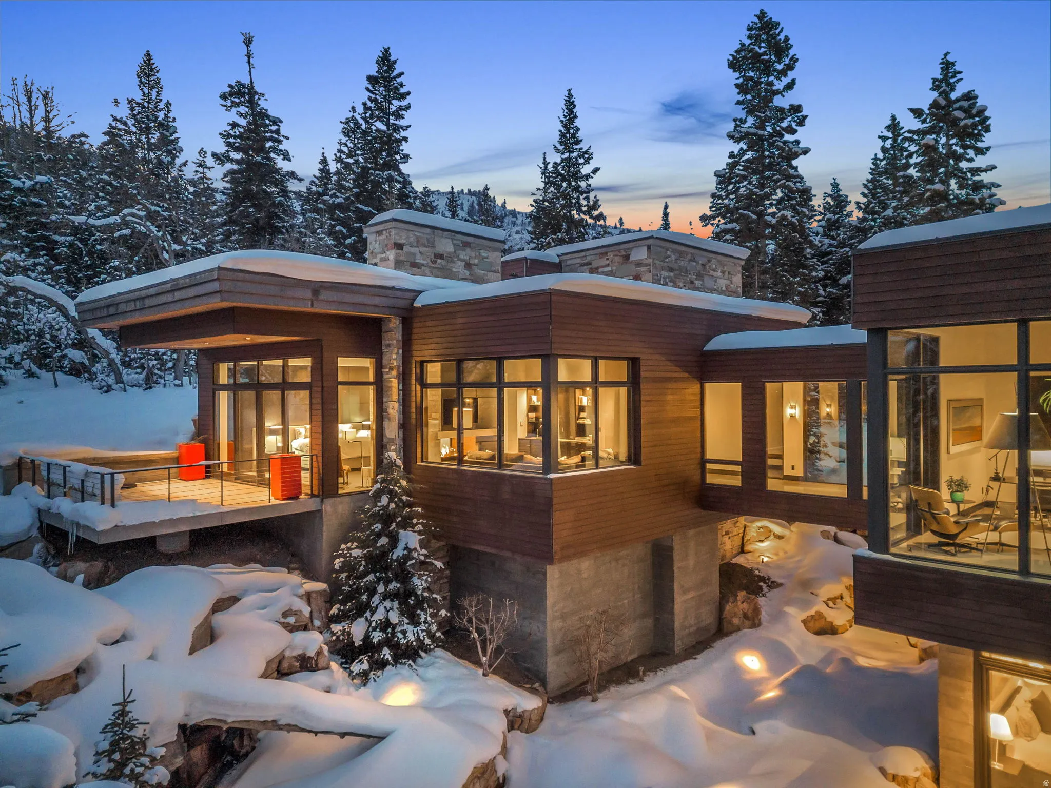 Modern mountain residence featuring extensive window walls, multi-level wood-clad exteriors, stone masonry accents, a suspended walkway, and a concrete foundational base