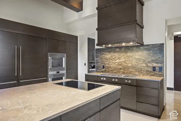 Kitchen featuring a large island with an integrated cooktop, dark wood-finish cabinetry, built-in stainless steel appliances, a stone backsplash, and a custom range hood