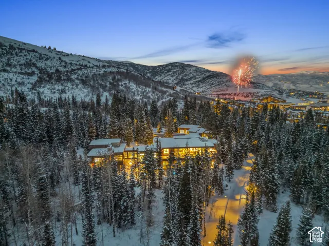 Extensive property nestled among snow-covered evergreen trees, featuring multiple levels, numerous illuminated windows, and a multi-peaked roofline
