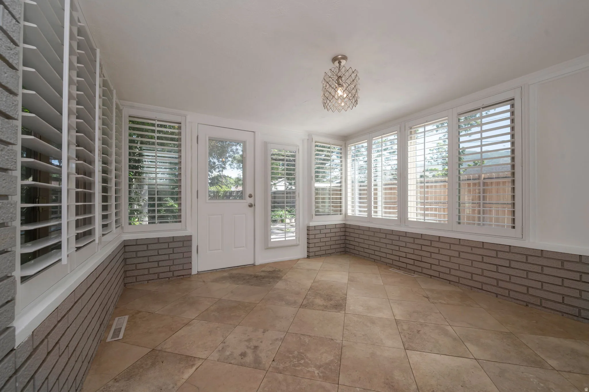 Bedroom 3/Den/Sunroom featuring diagonal tile flooring, a half-wall of brick, multiple windows with plantation shutters, a glass-paned exterior door, and a decorative ceiling light fixture