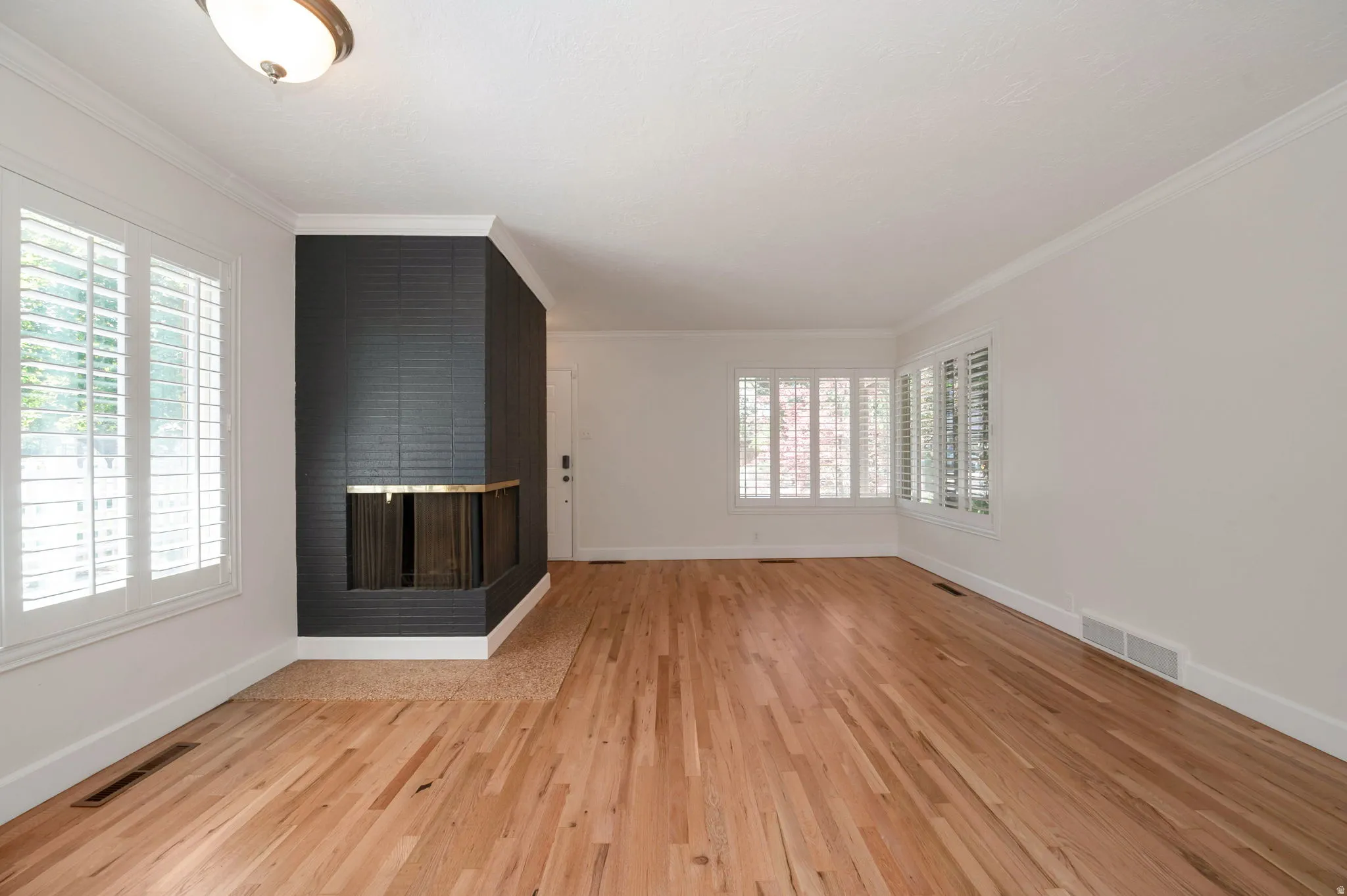 Expansive living area featuring hardwood flooring, a prominent brick fireplace with a brass accent, white crown molding, and multiple windows with white plantation shutters