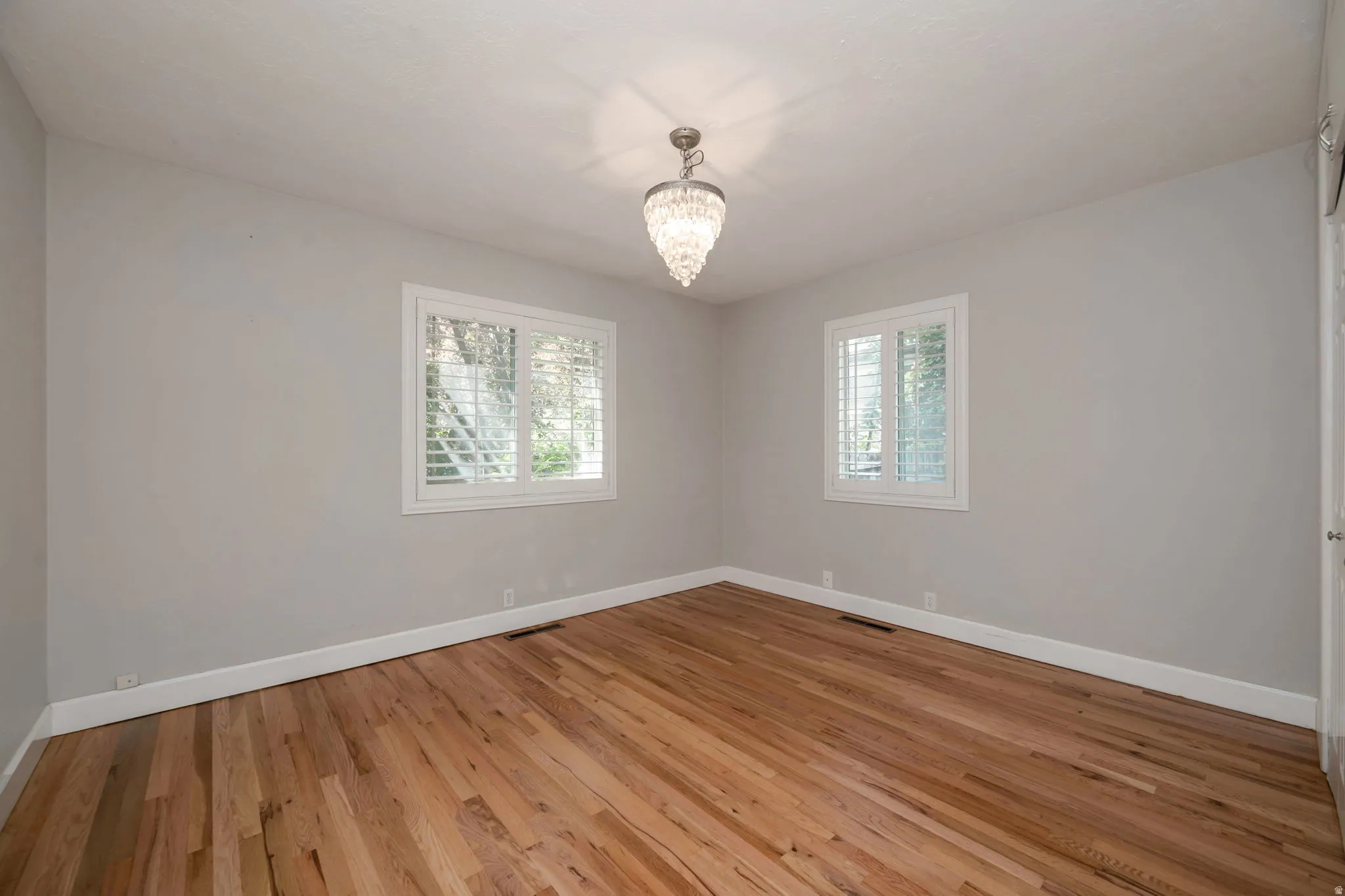 Bedroom 1 featuring wood-finish flooring, two windows with white plantation shutters, white baseboards, and a crystal chandelier