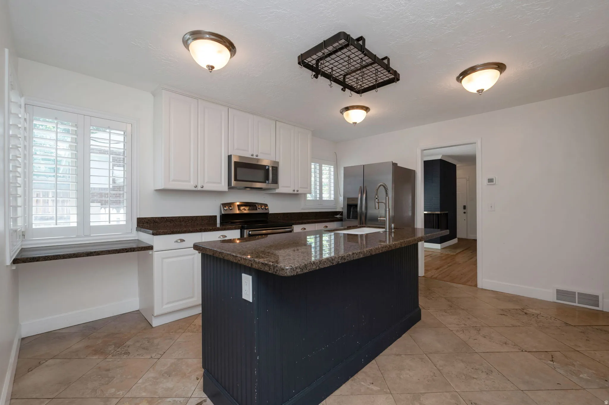 Kitchen featuring a dark-toned central island with a granite-style countertop and integrated sink, white upper and lower cabinetry, stainless steel appliances, and ceiling-mounted flush light fixtures
