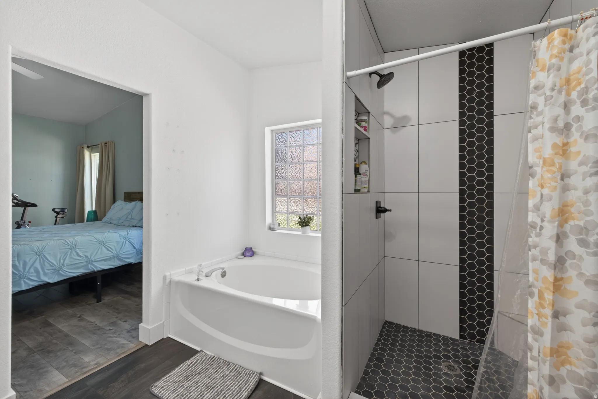 Bathroom featuring a corner soaking tub with a glass block window, dark wood-finish flooring, a shower with white subway tile, a dark hexagon tile accent strip, and a dark hexagon tile shower floor