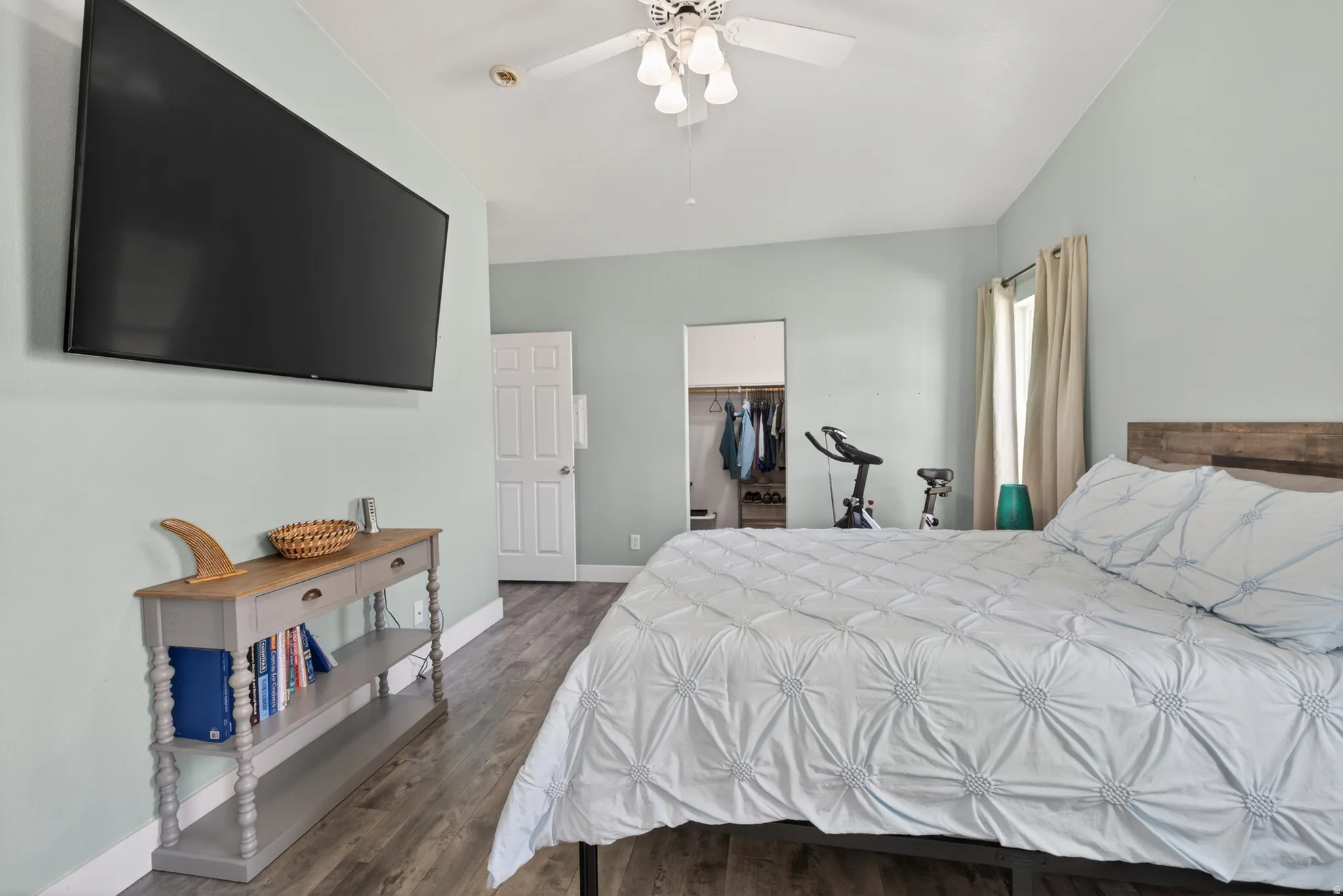 Room featuring wood-finish flooring, light blue wall paint, a built-in closet with shelving and hanging space, a window with curtains, and a ceiling fan