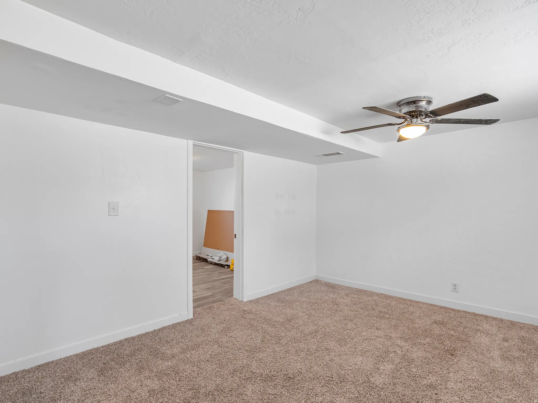 Carpeted room with white walls, a ceiling fan with integrated lighting, and an open doorway leading to a space with wood-finish flooring