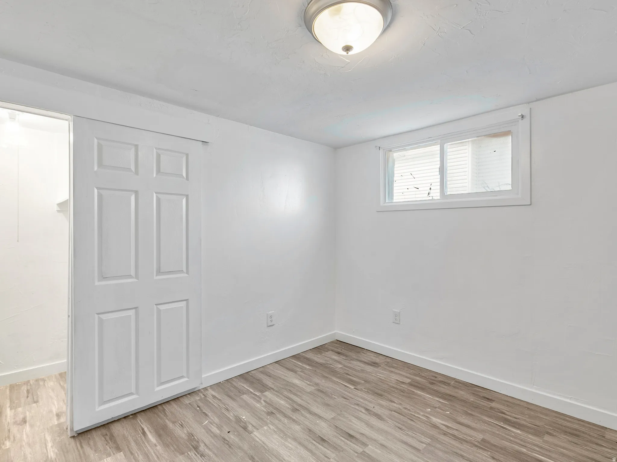 Room featuring wood-finish flooring, white walls, a single window with blinds, a ceiling-mounted light fixture, and a white paneled sliding barn door