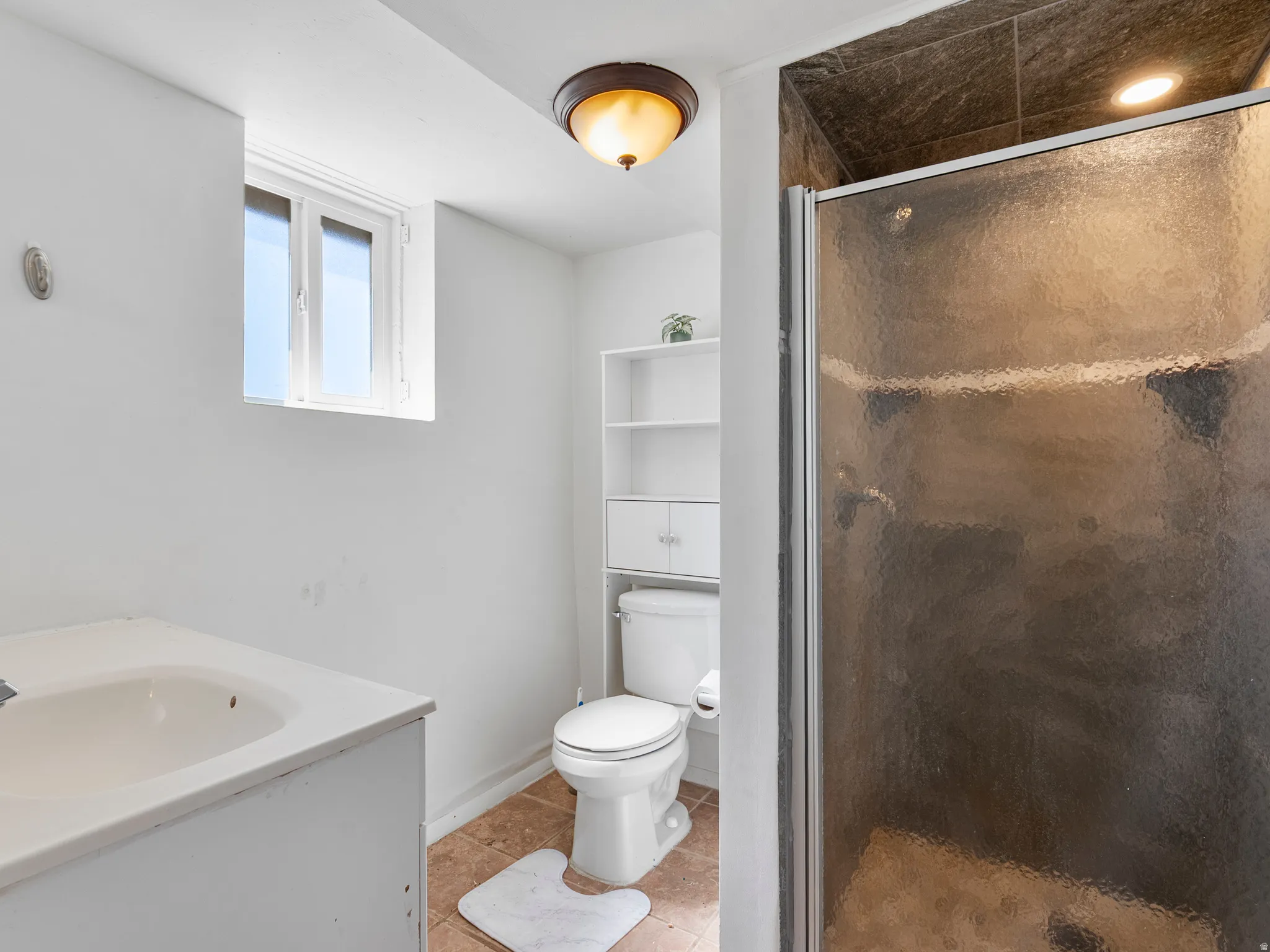 Bathroom featuring a tiled shower with a glass door, a vanity with an integrated sink, a commode, a built-in shelving unit, and a window