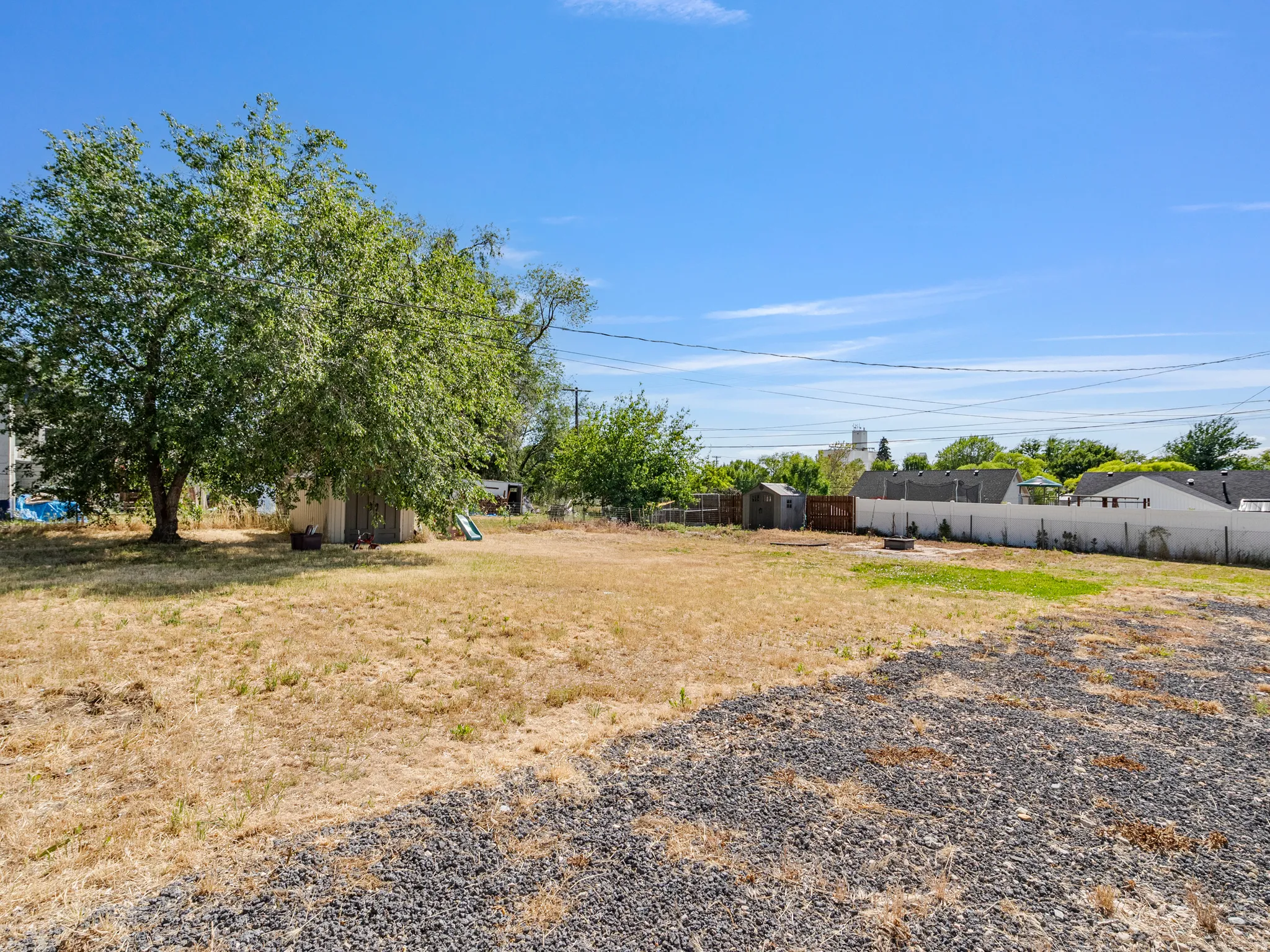 Expansive gravel and grass lot featuring a mature shade tree, a storage shed, and perimeter fencing