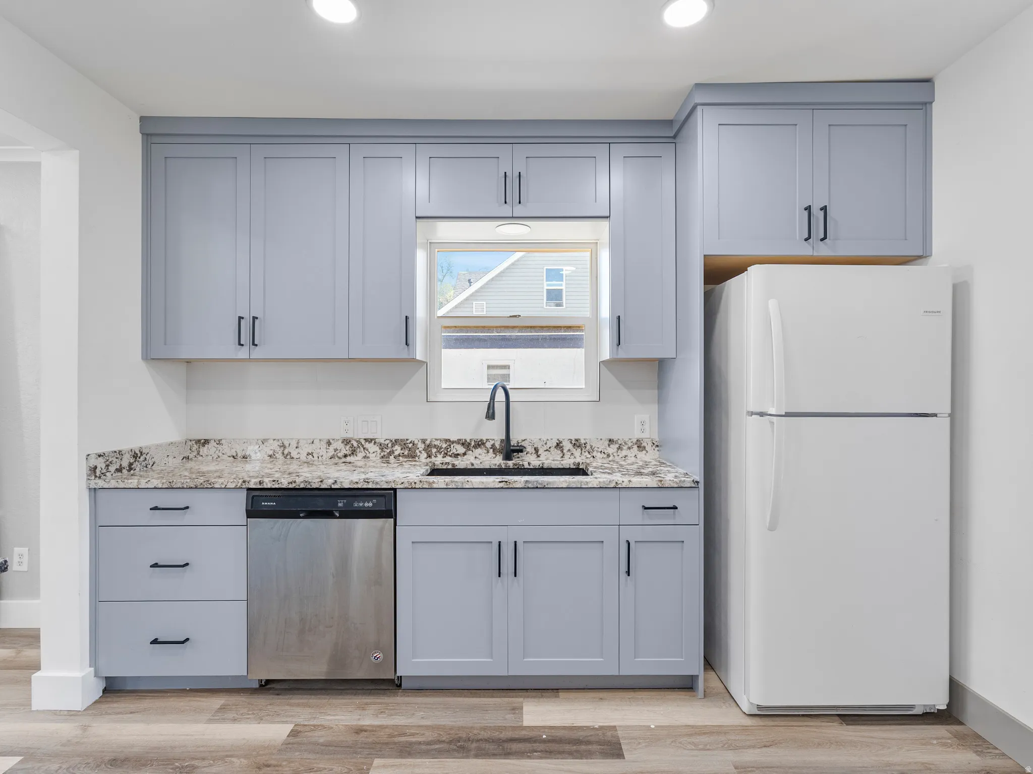 Contemporary kitchen featuring light gray cabinetry with black hardware, stone-finish countertops, stainless steel dishwasher, and wood-finish flooring